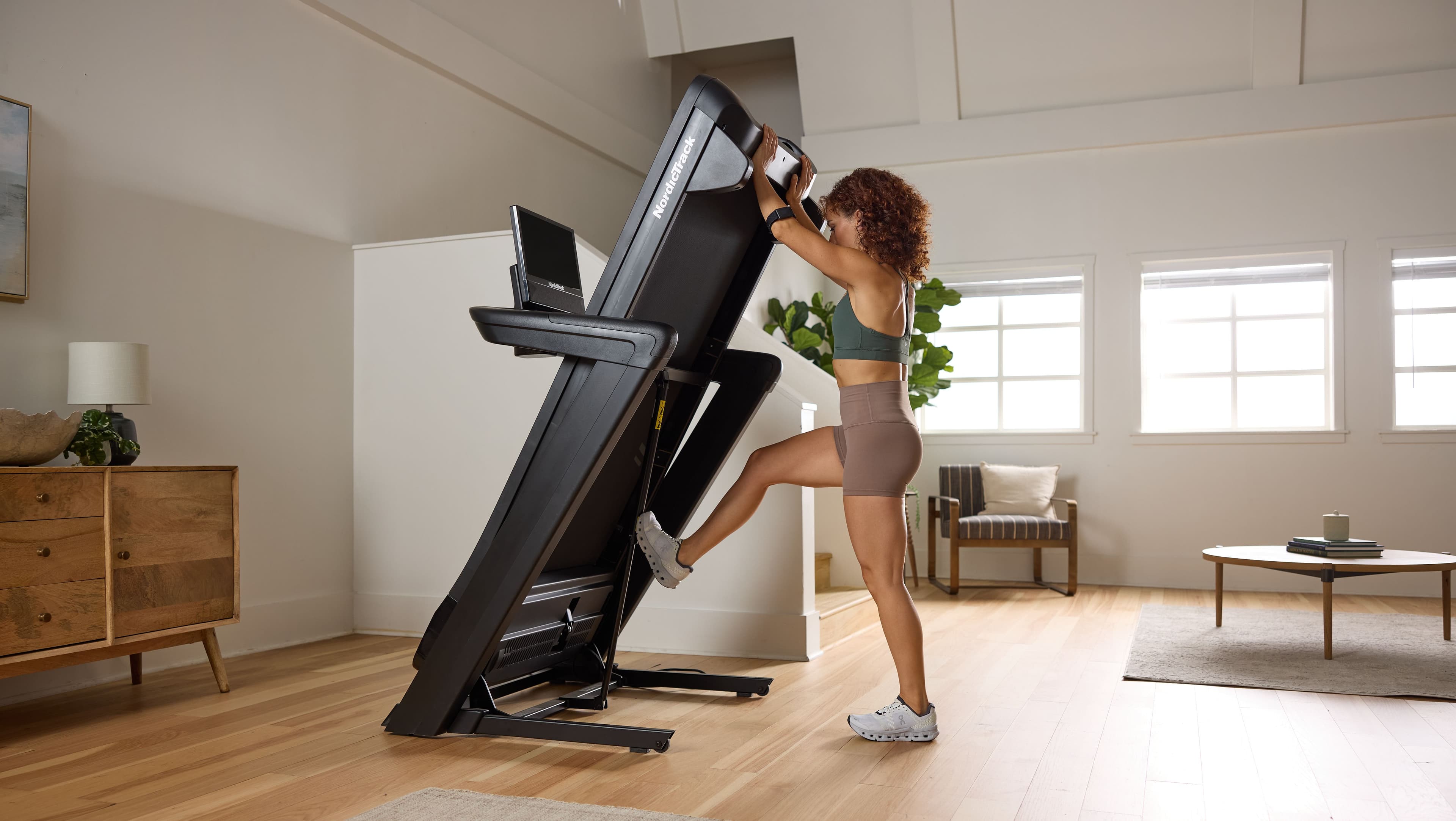 Woman folding a NordicTrack SpaceSaver treadmill upright in a bright apartment living room — showing compact storage for home use on hardwood floors.