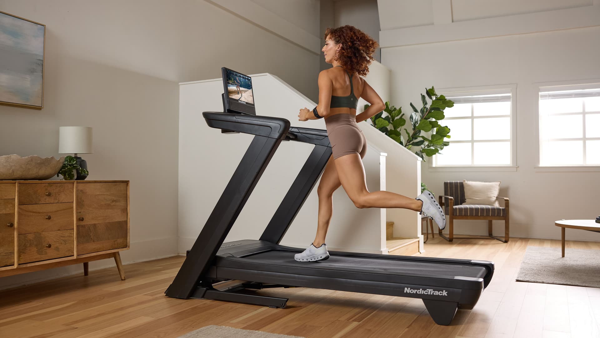 Lifestyle image showing a runner using a NordicTrack treadmill in a modern home gym environment while following an interactive workout on the touchscreen display.