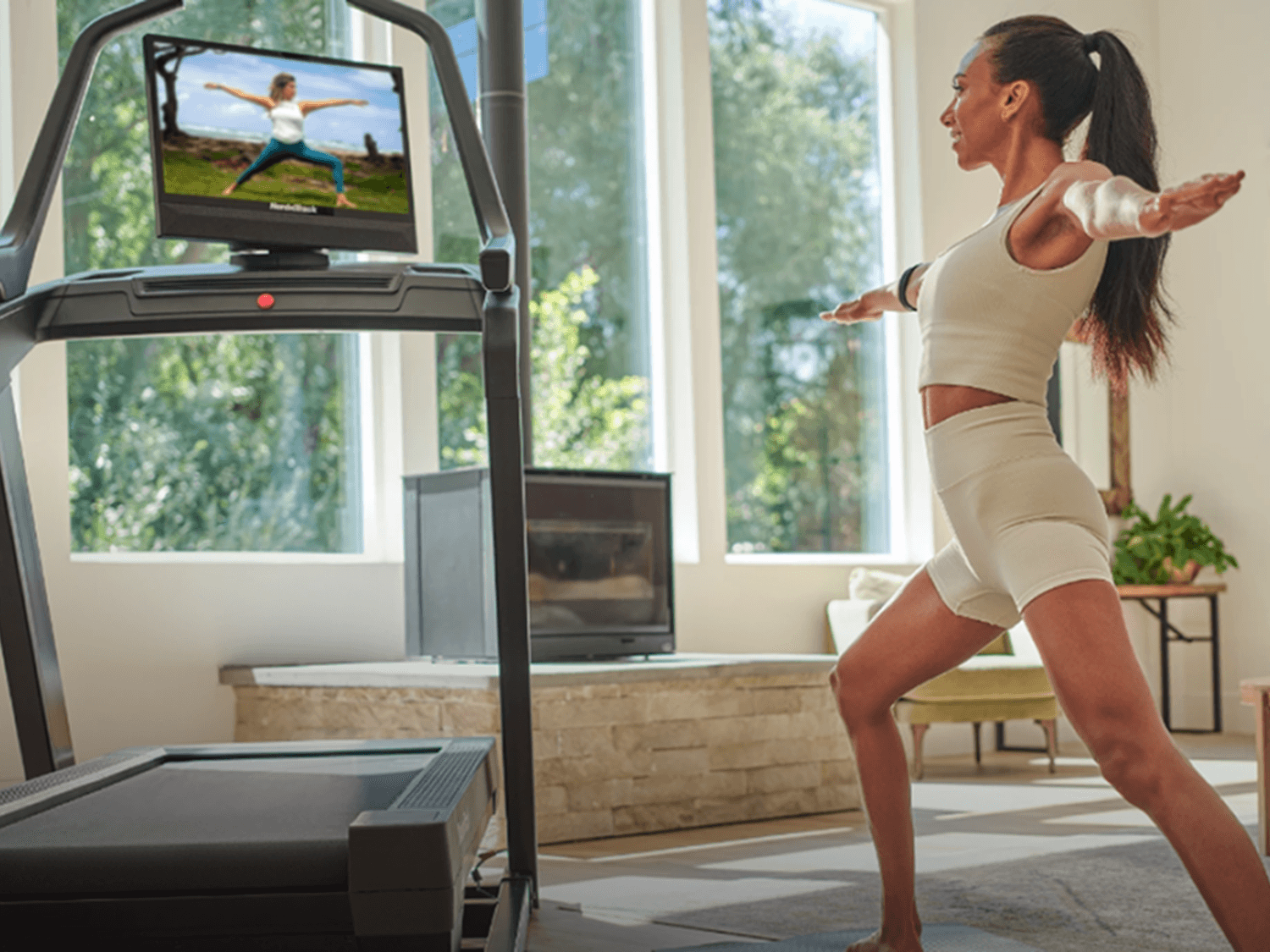 In a beautiful home with large windows, a woman stands next to her treadmill doing yoga, following a trainer on the rotated treadmill screen.